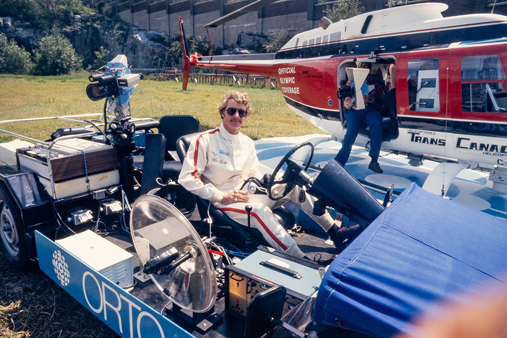 Un homme en combinaison blanche ornée d’un liseré rouge est assis à bord d’une autocaméra de l’Organisme de radiotélévision des Olympiques. Le véhicule bleu décapotable comporte de nombreux équipements, dont une caméra et une antenne circulaire.
