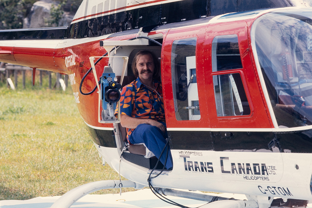 Un homme sourit, assis à bord d’un hélicoptère rouge à l’arrêt. Posant à côté d’une caméra, il porte la chemise de l’Organisme de radiotélévision des Olympiques. Le logo rouge, orange et jaune de Radio-Canada s’y déploie sur fond bleu marine.
