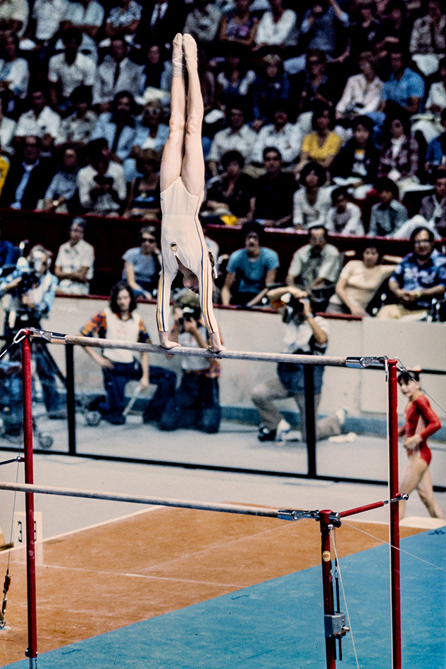 La jeune gymnaste Nadia Comăneci se tient en appui tendu renversé aux barres asymétriques. Elle porte un justaucorps blanc orné d’un liseré bleu, jaune et rouge devant la foule, deux photographes et un cameraman.