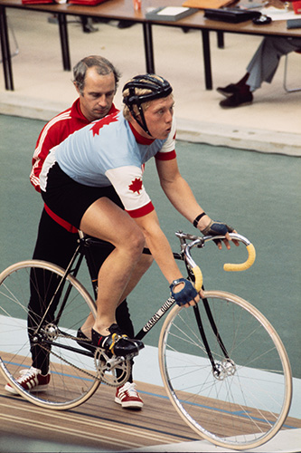 Le jeune cycliste sur piste canadien Gordon Singleton se tient à l’arrêt sur son vélo au cadre noir. Concentré, il expire tandis qu’un membre de son équipe l’aide à tenir en équilibre lors des préparatifs.