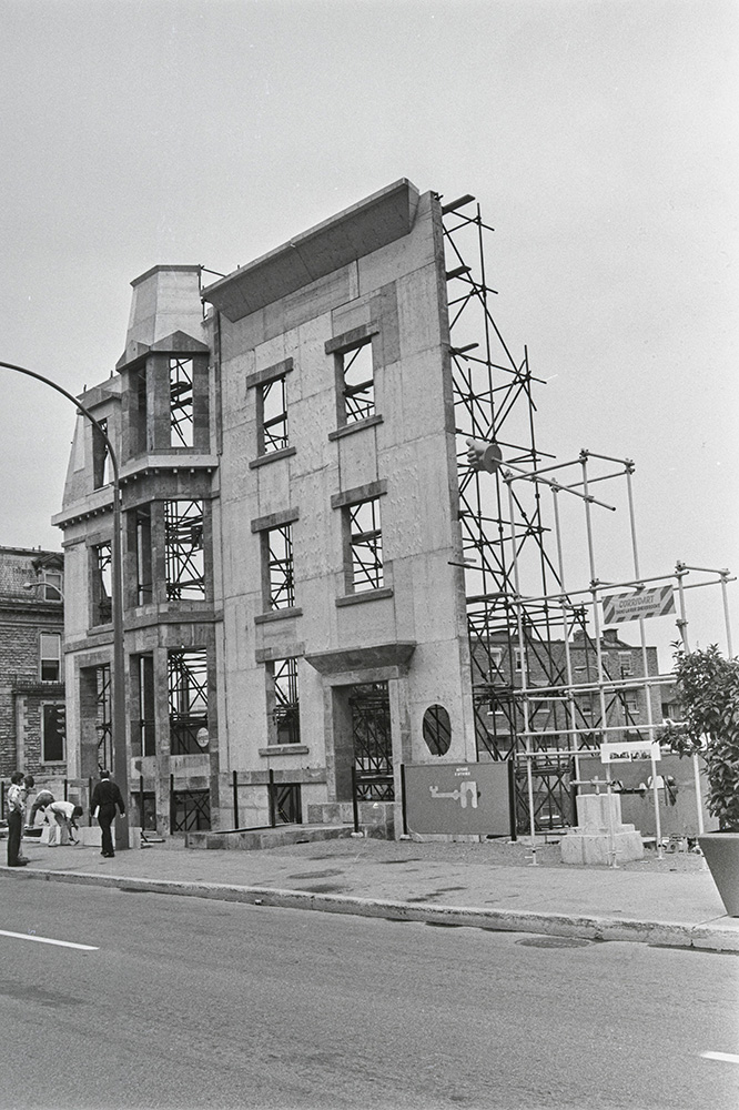 En noir et blanc, l'oeuvre d'art public intitulée <em>Les maisons de la rue Sherbrooke</em> de l'artiste Melvin Charney se dresse dans Montréal. Deux fausses façades imitent les résidences historiques du quartier, devant de hauts échafaudages.