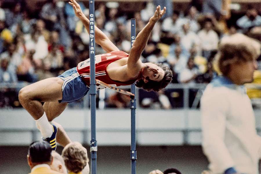 Caitlyn Jenner, à l'époque Bruce Jenner, réalise un saut en hauteur. Le dos cambré et les bras en extension, l’athlète en short bleu et maillot rouge passe tout juste au-dessus de la barre.