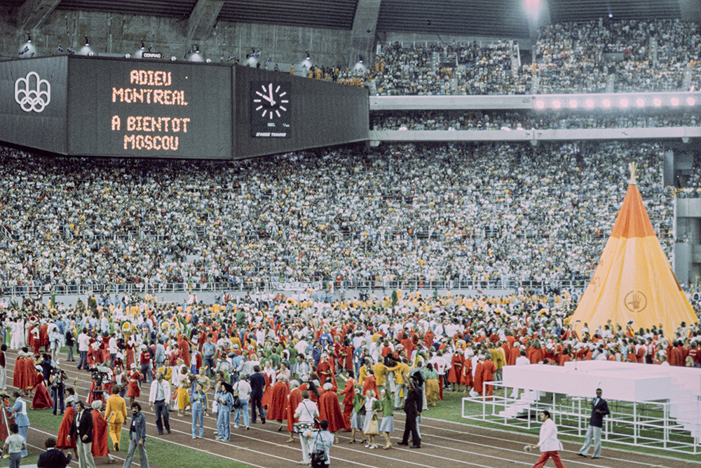 In the centre of the Olympic Stadium, a crowd of brightly costumed people wanders around a tall yellow teepee during the closing ceremony of the Games. In the background above the packed grandstands, a giant screen displays in French, "Farewell, Montreal, til we meet in Moscow."