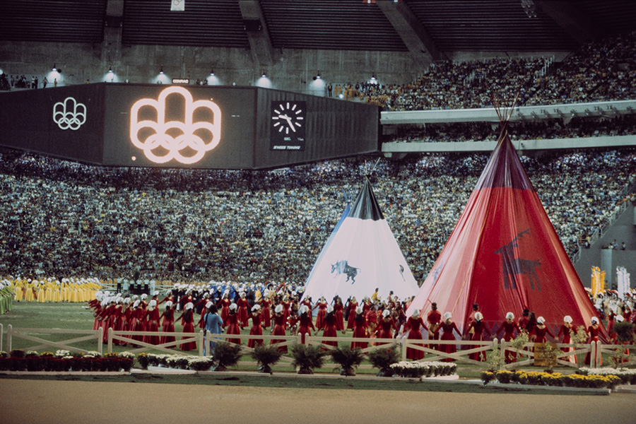 In the centre of the packed Montreal Olympic Stadium stand two teepees, one red, the other white. Both feature a stylized picture of an animal: a deer or caribou on the red one, and a buffalo on the white one. Many people dance around them, wearing outfits in solid colours.