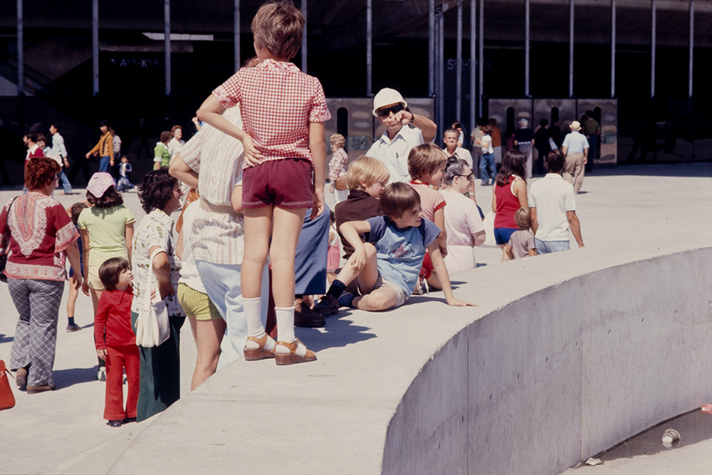 De nombreuses personnes, dont plusieurs enfants, marchent ou se tiennent devant le Stade olympique. Ils portent des habits d’été colorés.
