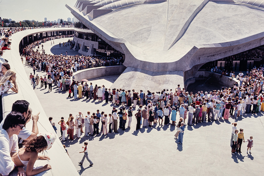 Devant le Stade olympique, une immense foule est réunie. Elle forme des files tout autour de la grande structure en béton gris clair du bâtiment.