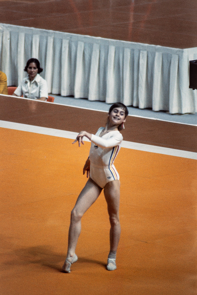 La jeune gymnaste Nadia Comăneci sourit lors d’une performance au sol, en justaucorps blanc orné d’un liseré bleu, jaune et rouge. Debout sur une surface orange, elle tend un bras vers l’avant et forme une pointe avec son pied opposé. Une femme assise la regarde.