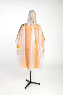 A mannequin is dressed in a clear hooded rain poncho, with a rainbow band running down the sleeves. Underneath the poncho can be seen a hospitality uniform from the Games: a bright orange midi skirt and matching jacket open to a white shirt.