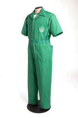 A mannequin is dressed in the maintenance staff uniform: an emerald green coverall with short sleeves, buttoned over a white t-shirt with a green neckline. The white Games logo is embroidered on the left chest pocket of the coverall.