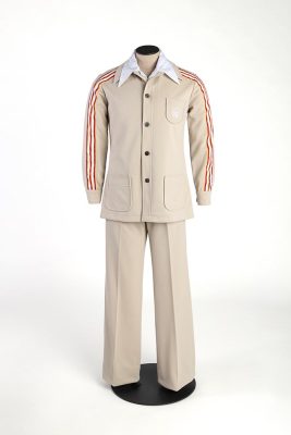 Un mannequin de couture porte l’uniforme du personnel auxiliaire : un pantalon droit beige et une veste de même couleur, boutonnée sur une chemise blanche. Un liseré tricolore orne les manches de la veste à la poche brodée du logo des Jeux.