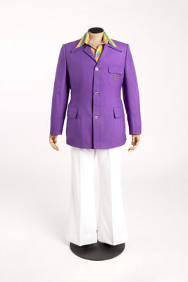 A mannequin is dressed in the medal presenter uniform: slightly flared white trousers and a multicoloured striped shirt under a purple jacket. A brooch of the Games logo is pinned to the breast pocket.