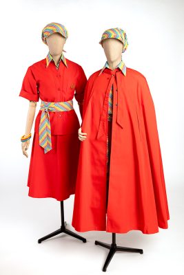 Deux mannequins de couture, coiffés d’un foulard à visière aux couleurs de l’arc-en-ciel, présentent l’uniforme d’hôtesse des Jeux. Celui de gauche présente une jupe mi-longue et une veste rouge vif à manches courtes, boutonnées sur une chemise au col rayé arc-en-ciel. Une ceinture reprend ce motif, tandis que des bracelets en plastique en déclinent les couleurs. Celui de droite présente une longue cape rouge vif laissant paraître une jupe et une veste de la même couleur, ouverte sur une chemise rayée multicolore.