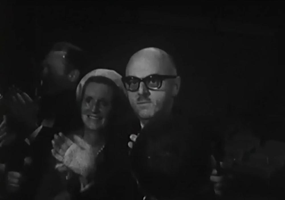 En noir et blanc, le maire Jean Drapeau se tient debout, accompagné de sa femme. D'un air surpris, il applaudit.