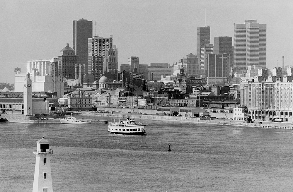En noir et blanc, les immeubles du Vieux-Port de Montréal et les gratte-ciel du centre-ville se dressent au bord du fleuve Saint-Laurent. Une navette fluviale s’éloigne du port.