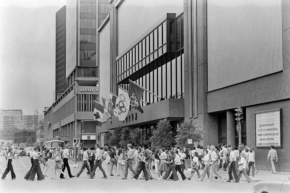 En noir et blanc, une foule traverse la rue Sainte-Catherine devant le Forum. Fixés à la façade de l’édifice, les drapeaux du Canada, du Québec et de Montréal flottent, comme ceux des Jeux olympiques et de leur édition montréalaise.