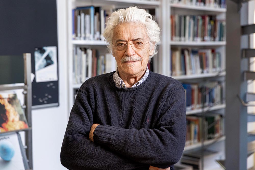Devant une bibliothèque, Raymond Bellemare se tient debout, les bras croisés. Vêtu d'un chandail foncé, il porte des lunettes.