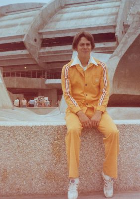 Devant le Stade olympique, un jeune homme assis sur un muret de béton sourit. Il porte l’uniforme du personnel d’accueil : un pantalon et une veste orange, aux manches ornées d’un liseré tricolore et à la poche brodée du logo des Jeux de 1976.