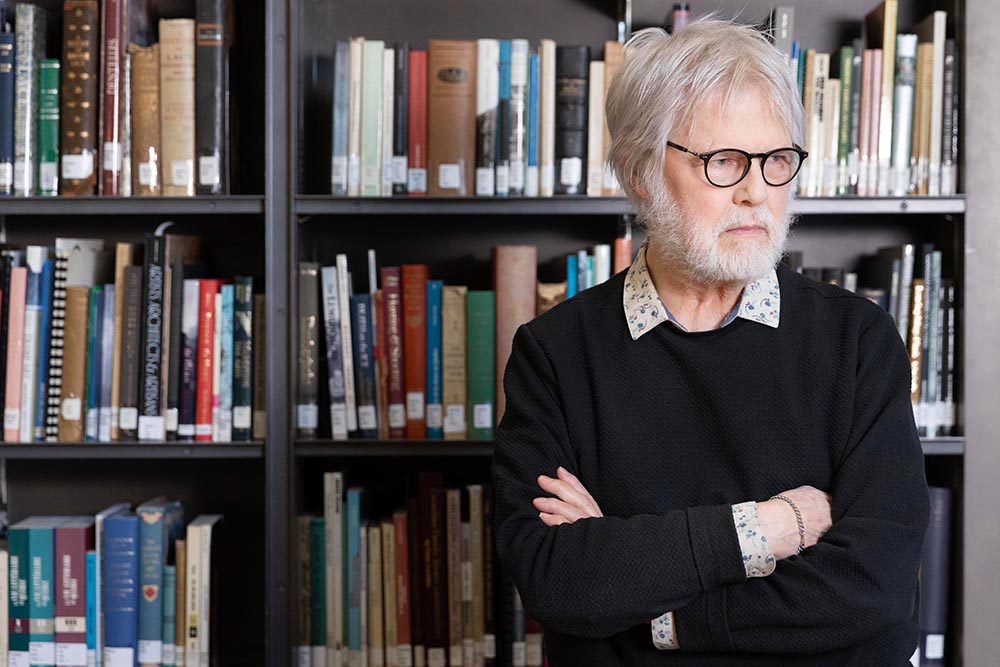 Devant une bibliothèque, Yvon Laroche se tient debout, les bras croisés. Vêtu d'un chandail foncé, il porte des lunettes. Son regard est tourné vers la droite.