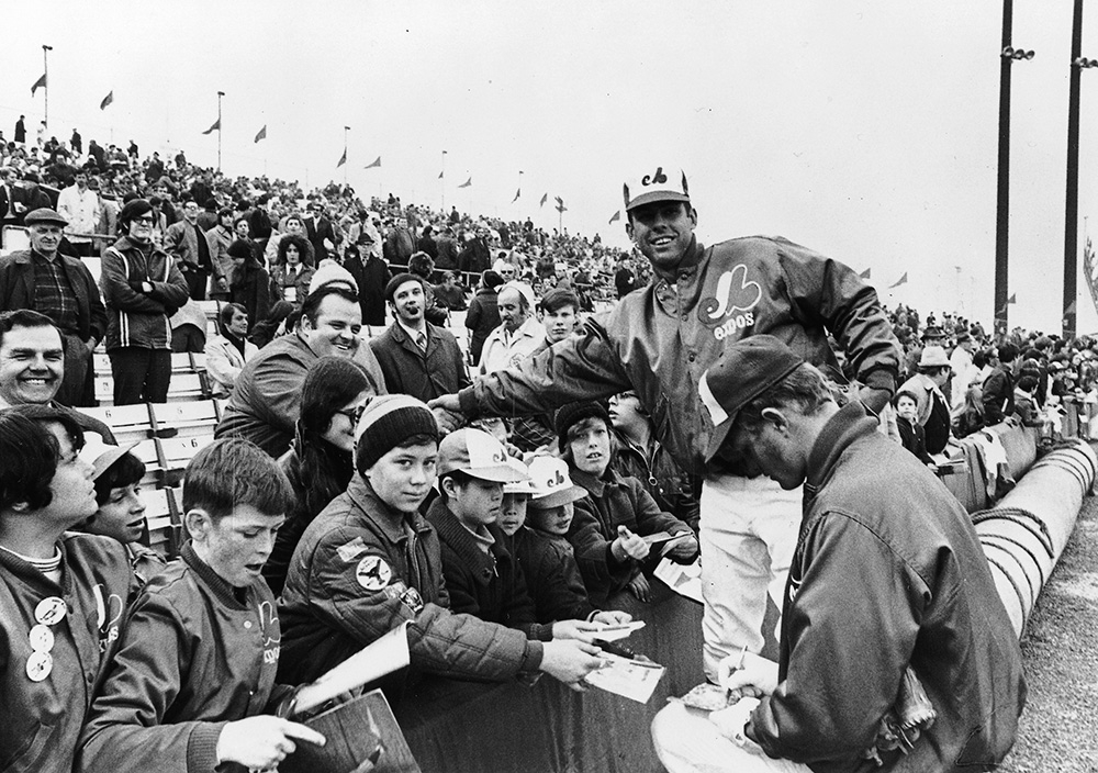 En noir et blanc, Ron Hunt, un joueur des Expos, serre la main d’un partisan. Au bas de gradins bien remplis, son coéquipier signe un autographe pendant que des garçons attendent leur tour, calepin à la main.