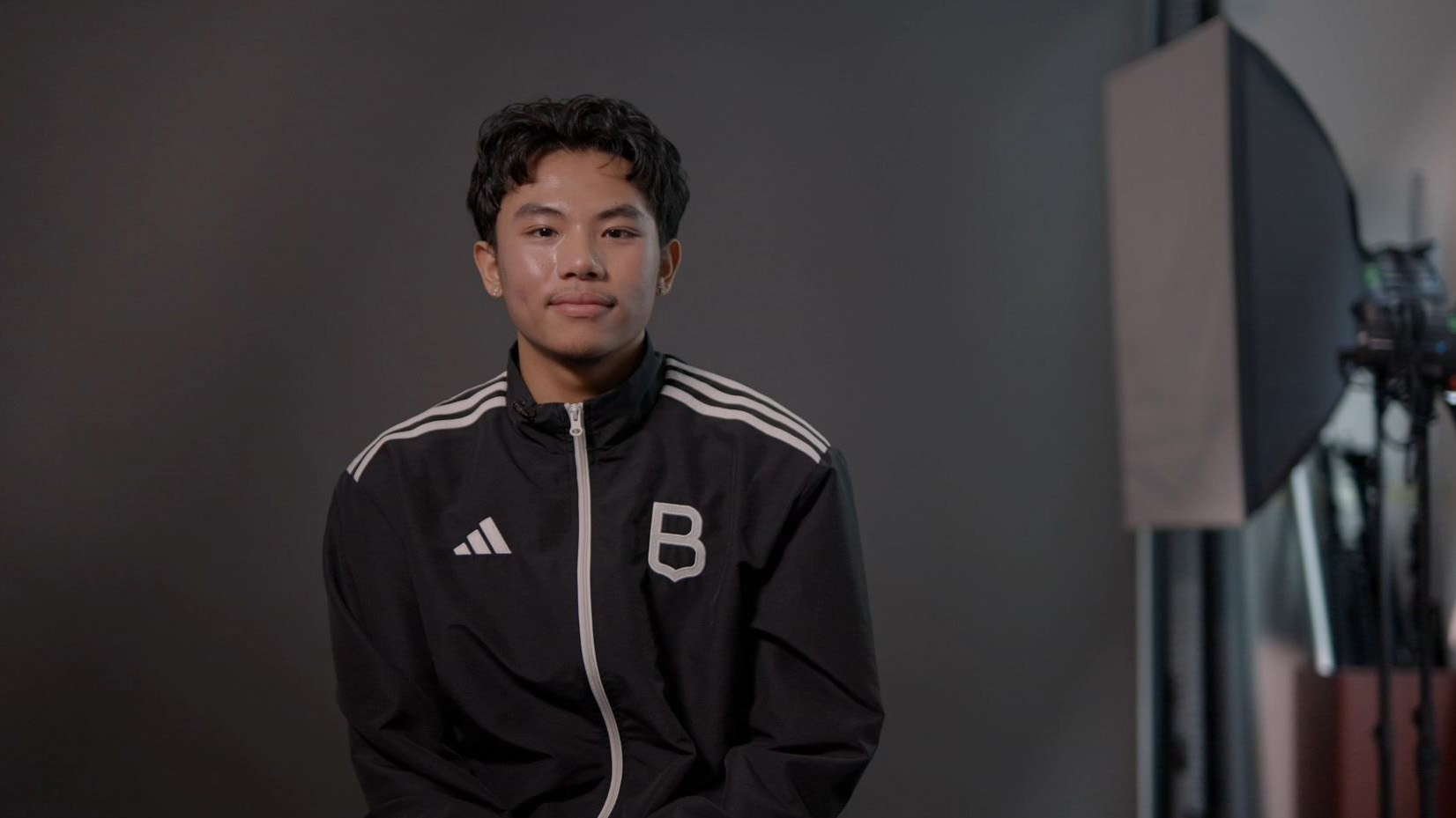 Moyen plan sur Aven Jairho Allana, souriant, devant un fond gris. Il porte une veste de sport noir à rayures blanches sur les épaules. Le logo adidas à droite, la lettre B stylisée, pour Brébeuf, à gauche.