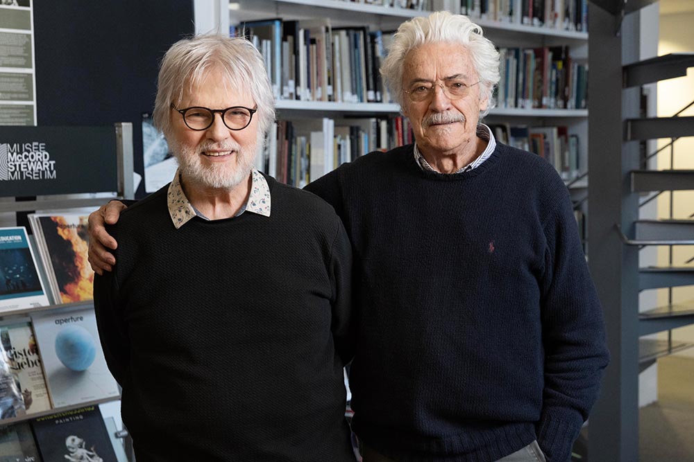 Devant une bibliothèque et un escalier à colimaçon, Yvon Laroche et Raymond Bellemare se tiennent debout, souriant. Les deux hommes sont vêtus d'un chandail foncé.