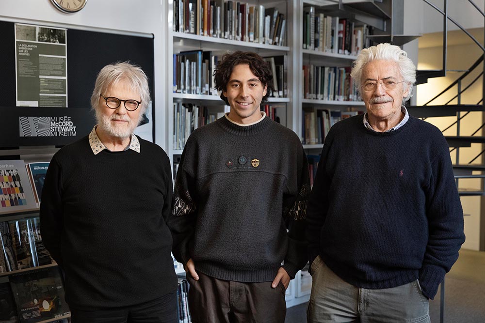 Devant une bibliothèque et un escalier en colimaçon, trois personnes se tiennent debout côte à côte. De gauche à droite : Yvon Laroche, Loïc Poisson et Raymond Bellemare. Les trois portent un chandail foncé et sourient.