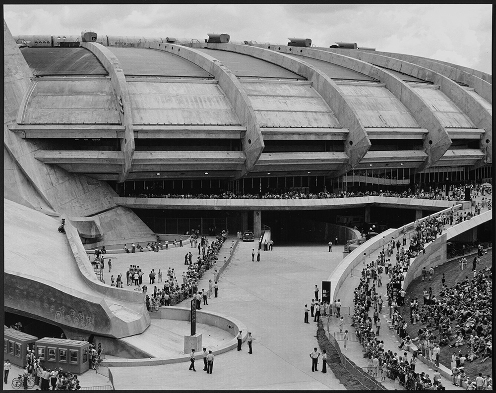En noir et blanc, l’immense base en béton du Stade olympique de Montréal rappelle la forme d’une soucoupe. Devant sa structure imposante, de nombreuses personnes se déplacent. D’autres se reposent dans une aire gazonnée.