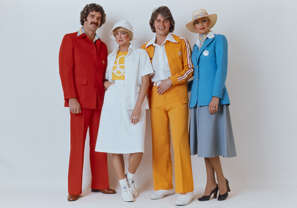 Quatre personnes présentent des uniformes des Jeux. Un homme porte un complet rouge. Une femme présente une tenue blanche décontractée à la jupe mi-longue. Un homme est vêtu d’un ensemble orange. Une femme porte un tailleur bleu clair à la veste cintrée.