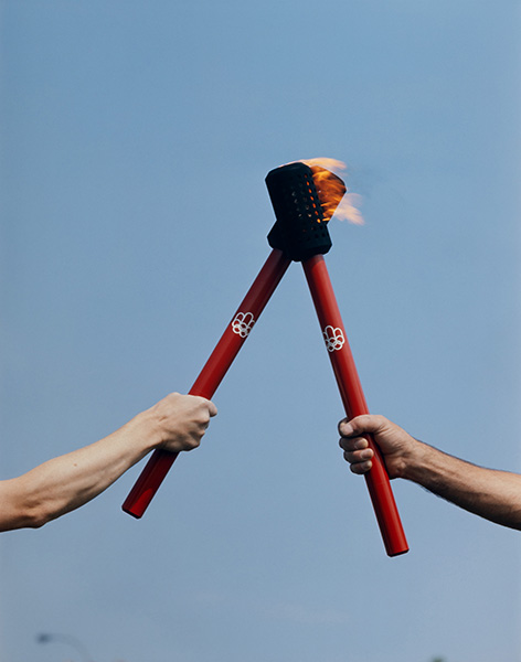 Two arms each brandish an official torch of the Montreal Olympic Games. The black upper part of one torch touches that of the other, passing on the Olympic flame. The red aluminium handles feature the Games logo printed in white.