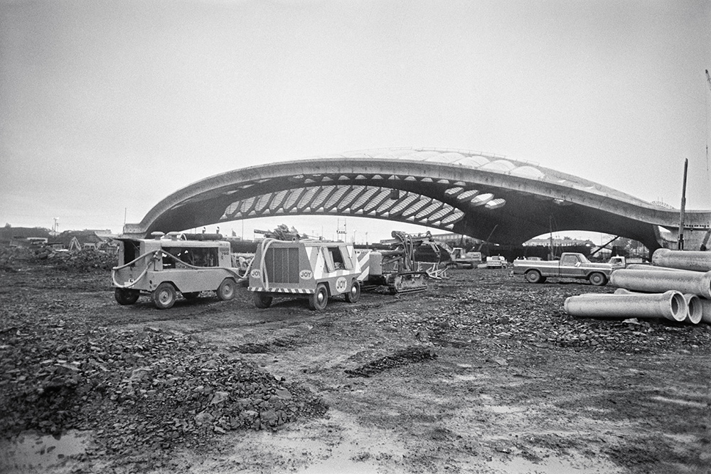 En noir et blanc, l’extérieur du Vélodrome en construction s’impose au milieu d’un vaste terrain boueux. Sa toiture ajourée se découpe sur le ciel clair et des véhicules de chantier ainsi que de gros conduits cylindriques occupent le premier plan.