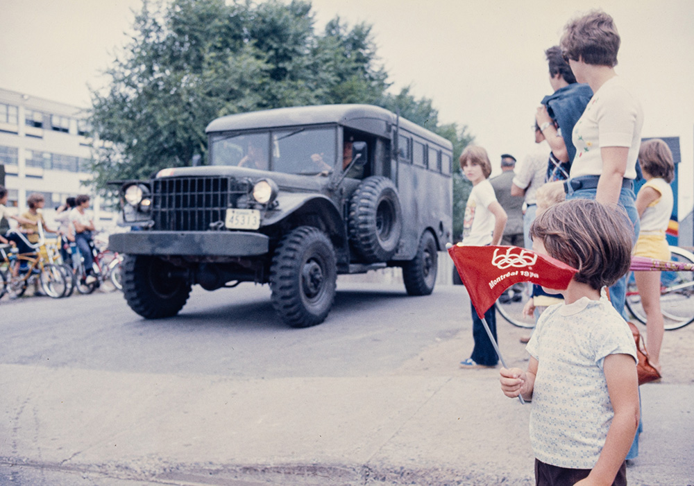 Dans une rue de Montréal, un camion militaire circule devant la foule. Un enfant secoue un petit drapeau rouge et blanc des Jeux olympiques. Une famille et des cyclistes à l'arrêt regardent aussi le camion passer.