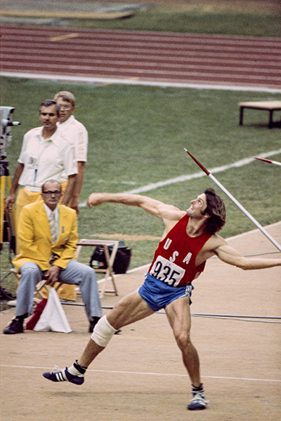Caitlyn Jenner, à l'époque Bruce Jenner, se tient en appui sur une jambe, sur le point de lancer son javelot. L’athlète porte un short bleu et un maillot rouge où il est écrit, USA, et le numéro 935. Trois arbitres habillés en jaune, bleu pâle et blanc l’observent en arrière-plan.