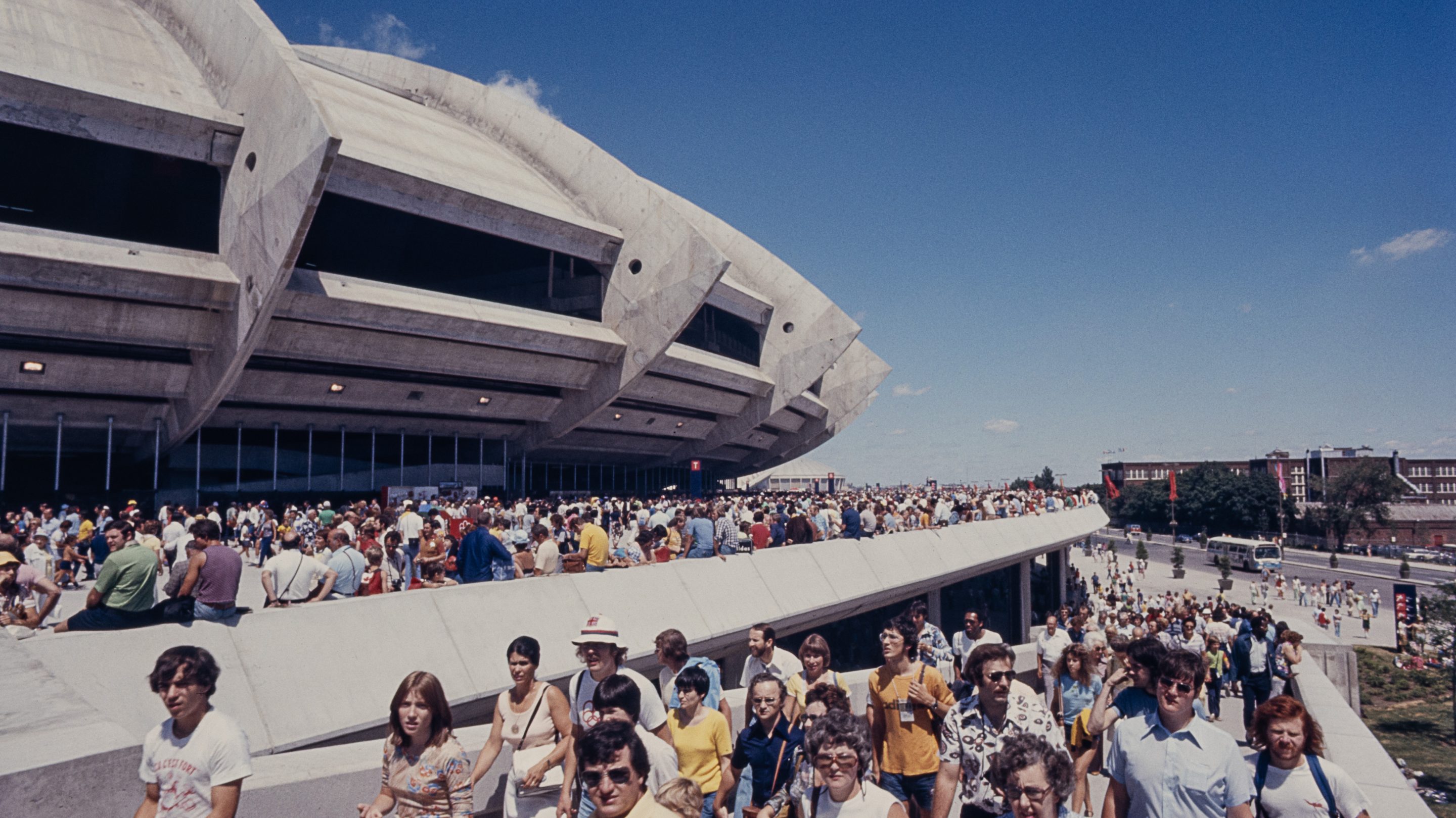 De nombreuses personnes vêtues d’habits d’été colorés se dirigent vers le Stade olympique, en empruntant une large rampe de béton. D’autres sont assises sur le rebord de l’esplanade bondée entourant le bâtiment.