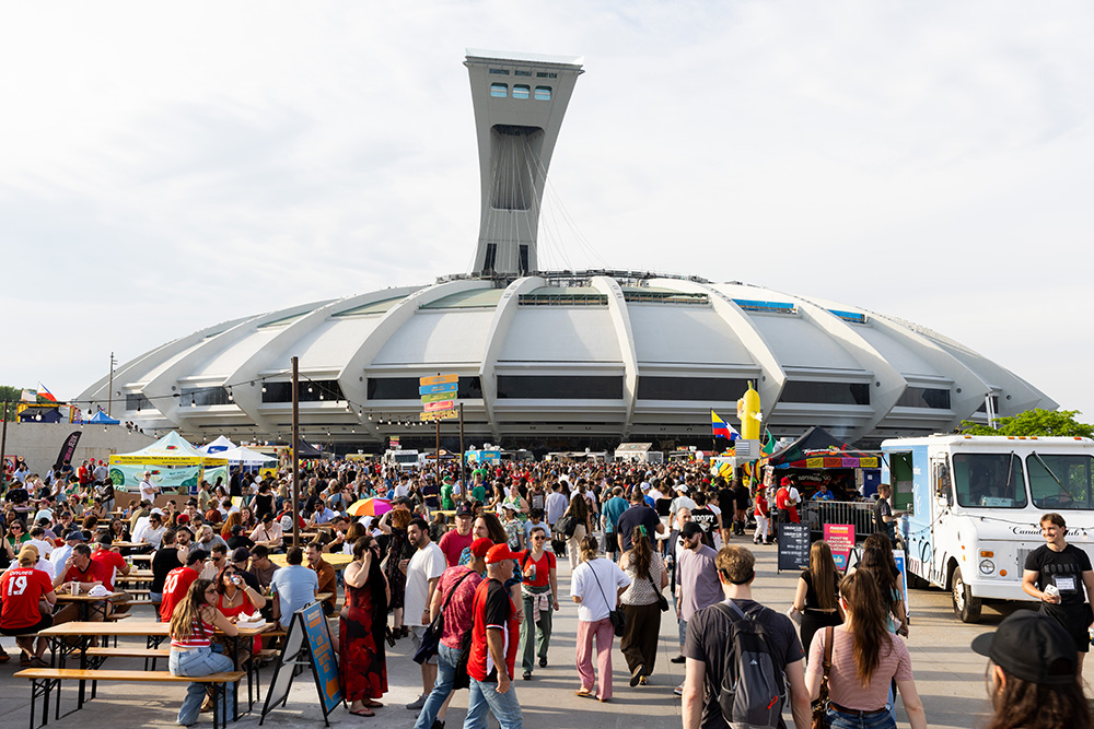Devant le Stade olympique, sur l’esplanade, une foule estivale est réunie. À gauche, des personnes sont assises autour de tables de pique-nique. D’autres se promènent près de camions de cuisine de rue dispersés sur le site.