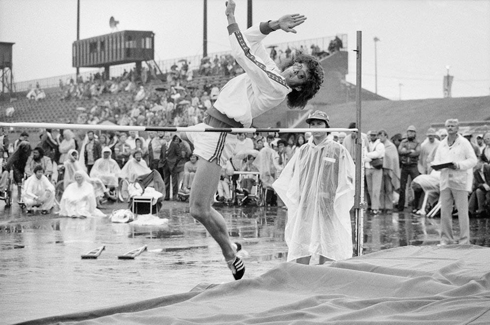 En noir et blanc, l’athlète paralympique canadien Arnold Boldt, amputé d’une jambe, réalise un saut en hauteur. Le dos cambré et les bras en extension, il amorce son passage au-dessus de la barre. L’arbitre et le public le regardent, sous leurs vêtements de pluie.