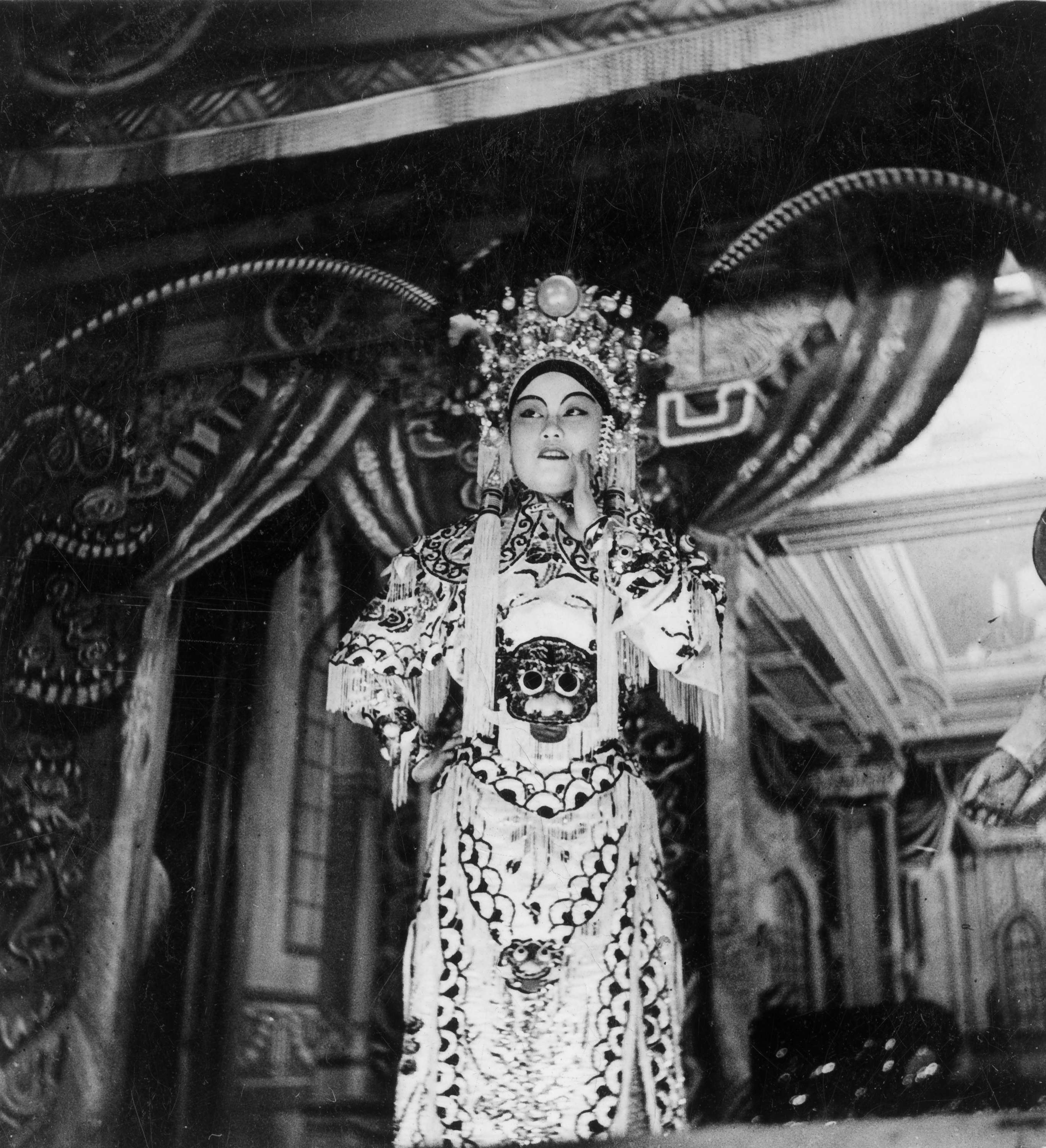 <span style="font-weight: 400;">Une actrice d’opéra cantonais se tient sur scène, regardant le public avec un sourire discret. Elle porte une coiffe ornée de longues pampilles et de plumes de faisan, ainsi qu’un costume de couleur claire dont le torse est décoré d’une tête de lion.</span>