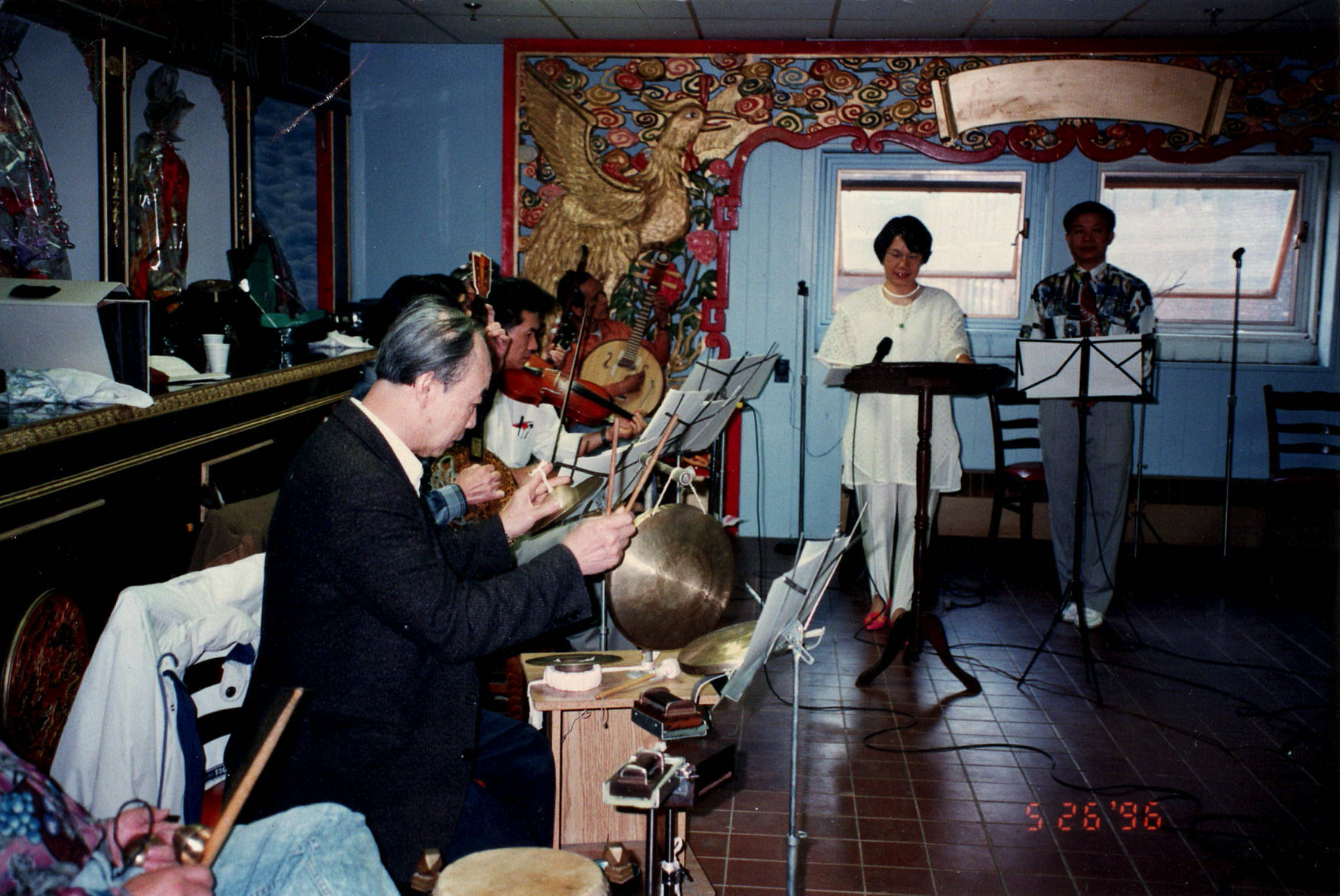 <span style="font-weight: 400;">Au premier plan à gauche, des personnes jouent de la musique lors d’une répétition, alors que deux interprètes les accompagnent au chant, plus loin dans la pièce. On aperçoit une peinture murale détaillée d’un phénix. La photo est datée du 26 mai 1996.</span>