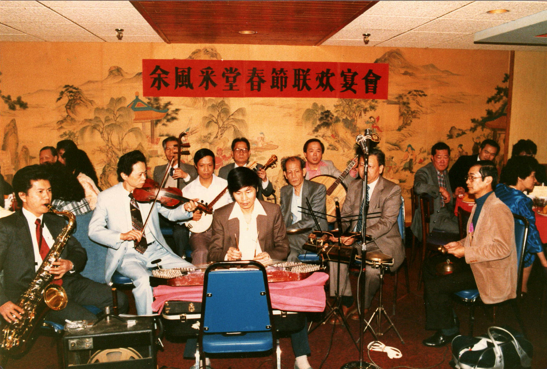<span style="font-weight: 400;">Un groupe de dix musiciens vêtus de costumes dans le style des années 1970 se produit dans un restaurant de Montréal avec divers instruments traditionnels et contemporains. En arrière du groupe, on aperçoit la peinture d’un paysage et une banderole rouge sur laquelle sont inscrits des caractères chinois.</span>