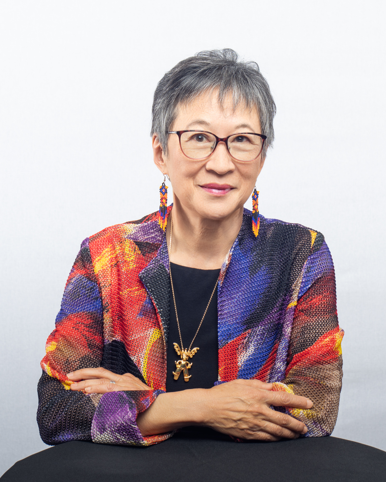 <span style="font-weight: 400;">Portrait de Julie Jai assise à une table, souriant doucement à la caméra. Elle porte un blazer coloré, des boucles d’oreilles en perles et des lunettes de vue.</span>