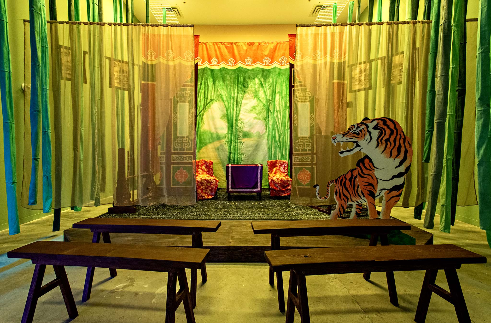 View of Scent of Thunderbolts from directly in front of the stage. Wooden benches are arranged for the audience, fabric strips resembling bamboo hang on both sides, and a painted tiger cutout appears at the right of the stage with chairs and drapes in the background.