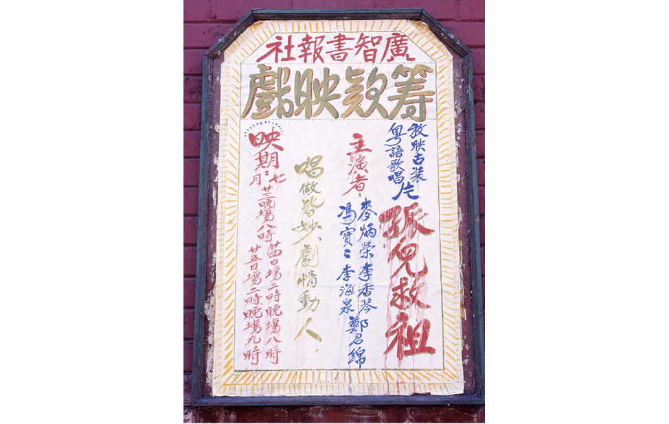 <span style="font-weight: 400;">Une affiche encadrée avec des caractères chinois aux couleurs vives écrits à la main est accrochée à l’extérieur sur un mur de briques rouges. Il y a des taches d’encre dans la partie inférieure.</span>