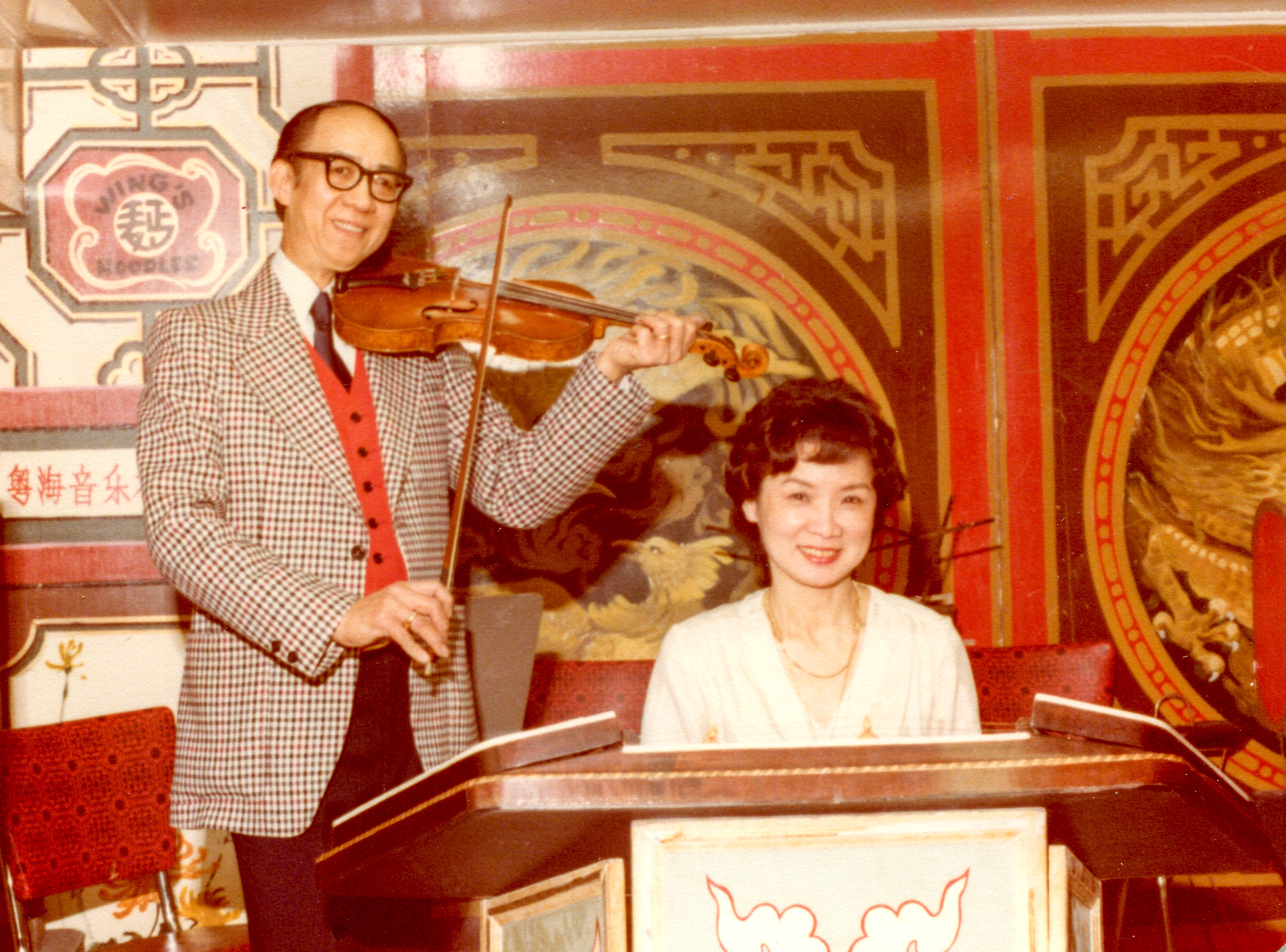 <span style="font-weight: 400;">Raymond Jai joue du violon tandis que Beatrice Jai est assise à ses côtés, regardant directement l’appareil photo.</span>