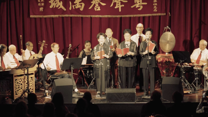 <span style="font-weight: 400;">Une scène pleine avec des chanteur·teuse·s debout au centre entouré·e·s de musicien·ne·s. Une banderole horizontale rouge avec du texte en caractères chinois est suspendue au-dessus de l’ensemble.</span>