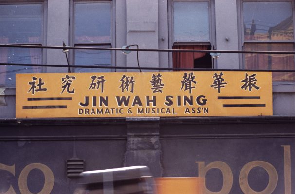 <span style="font-weight: 400;">An orange sign with black text in both Chinese and English for the Jin Wah Sing Dramatic & Musical Association.</span>