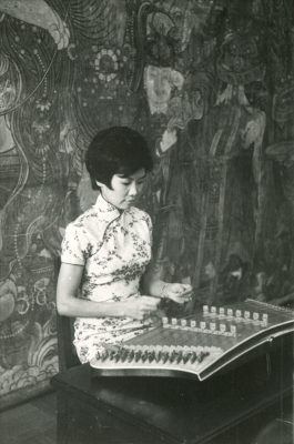 <span style="font-weight: 400;">黑白照片，廖瑤華女士坐在壁畫前用竹槌演奏揚琴。</span>