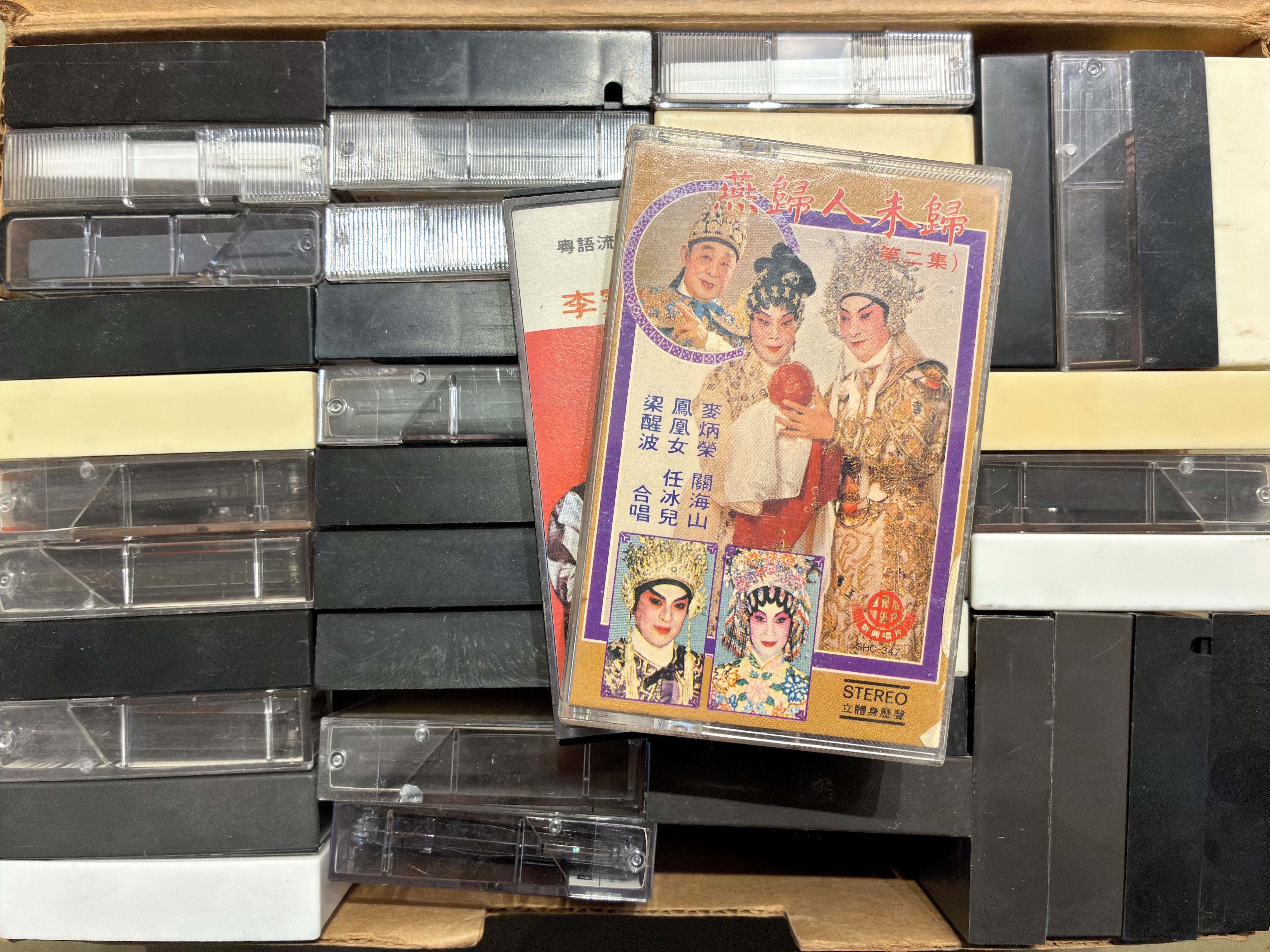 <span style="font-weight: 400;">Gros plan sur une boîte remplie de cassettes audio. Plusieurs cassettes sont empilées sur le dessus, laissant apparaître leurs pochettes illustrées. La pochette sur le dessus représente deux personnages principaux souriant à un enfant; les portraits plus petits d’autres artistes les entourent.</span>