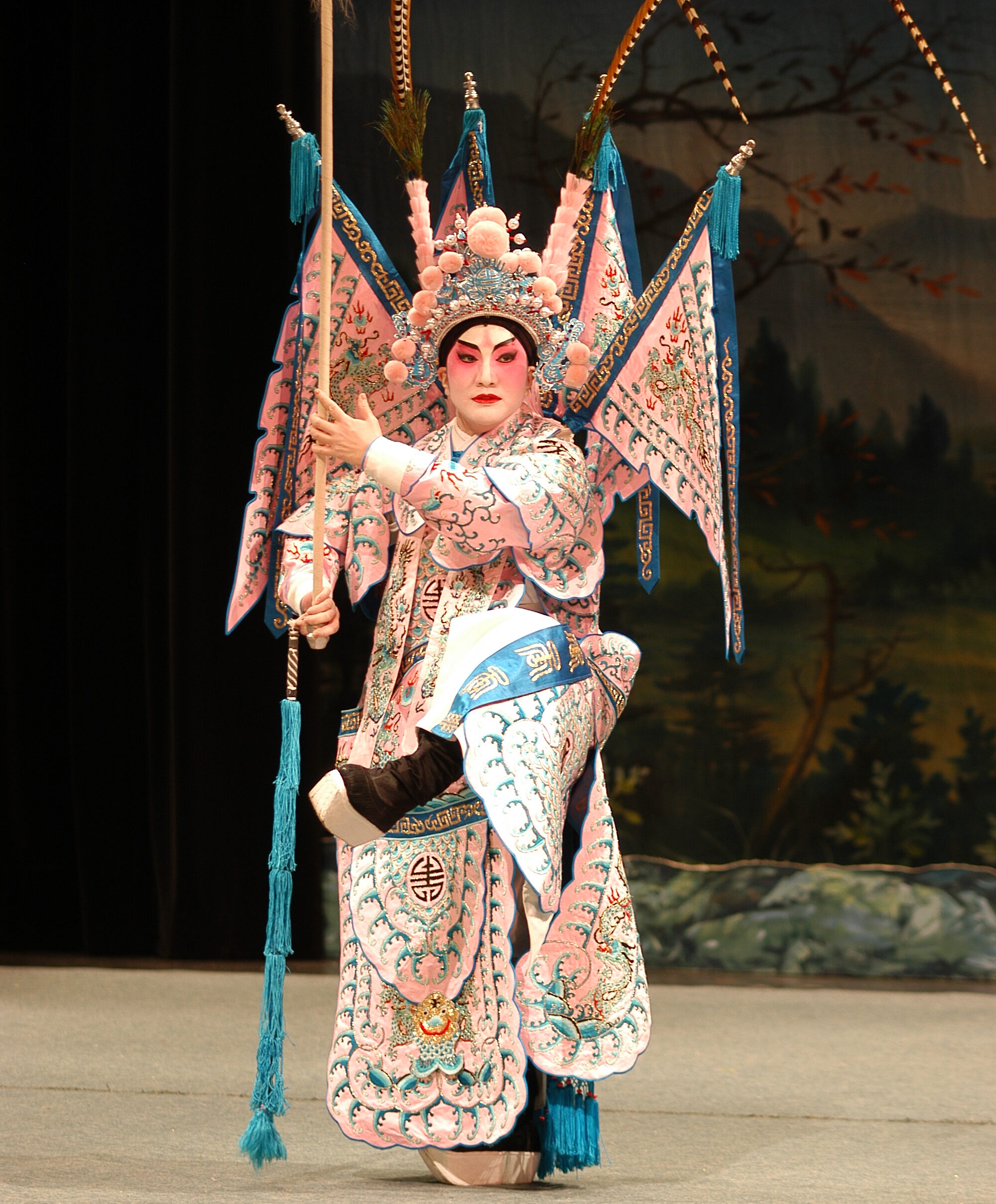 <span style="font-weight: 400;">Alice Chan performs as a high-ranking warrior, wearing back-mounted flags with long feathers rising from her headdress, poised to strike with a spear.</span>