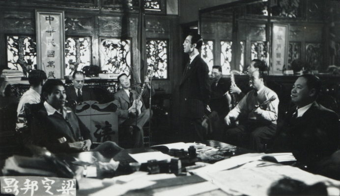 <span style="font-weight: 400;">Photo en noir et blanc montrant plusieurs hommes assis en cercle dans une salle de répétition, jouant de leurs instruments, tandis qu’un homme se tient debout au centre, chantant, les mains nonchalamment jointes derrière le dos.</span>