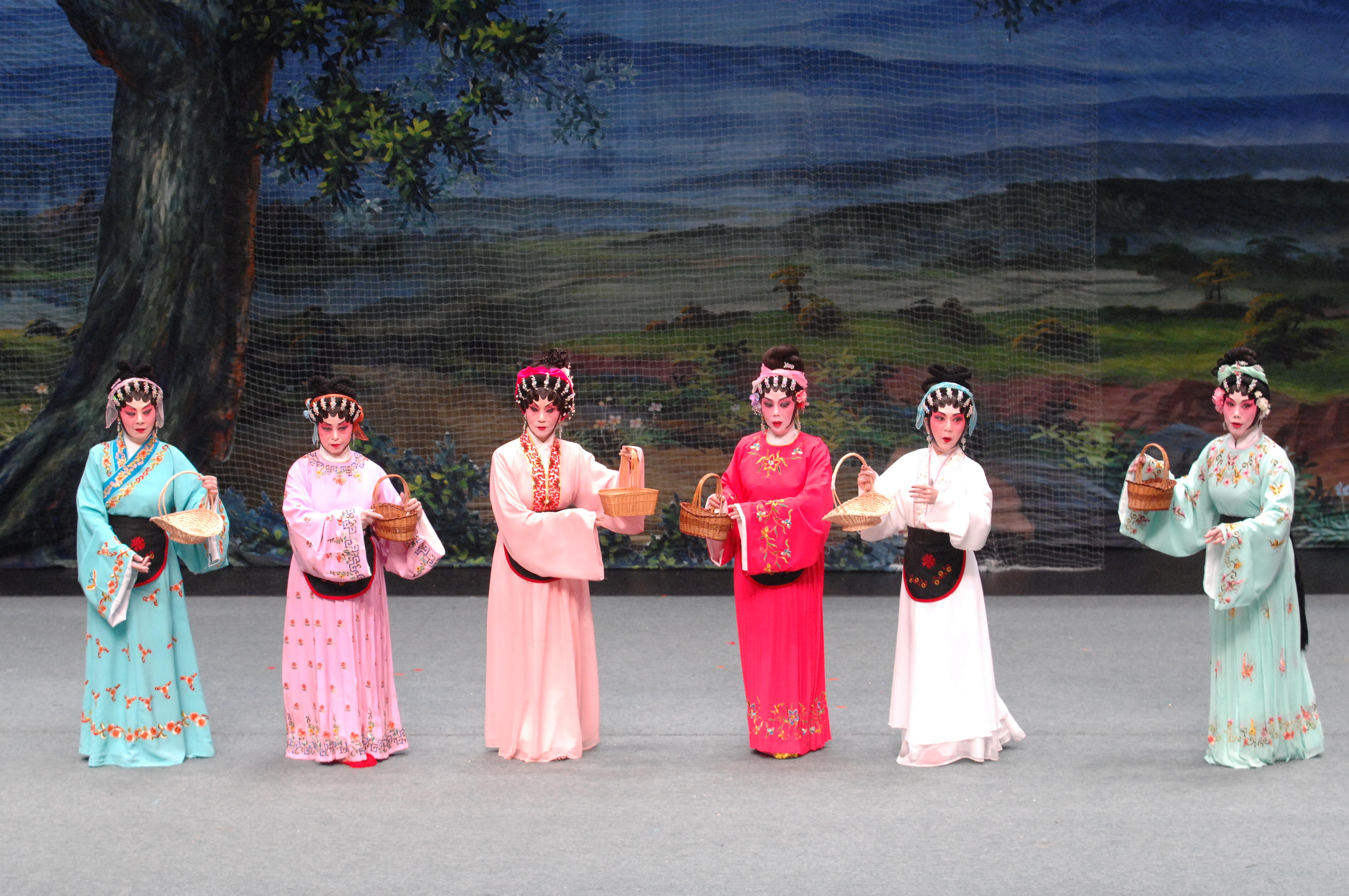 <span style="font-weight: 400;">Six actrices sont alignées devant une toile de fond dépeignant un paysage. Elles portent des coiffes et des tabliers assortis, chacune un panier à la main. Leurs costumes sont colorés et dotés de longues manches d’eau.</span>