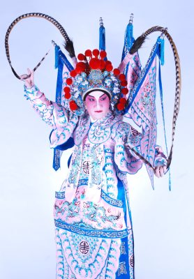 <span style="font-weight: 400;">Une artiste porte un costume élaboré rose et bleu orné de motifs de dragons avec des drapeaux fixés à son dos. La coiffe richement décorée est agrémentée de pompons rouges et de deux longues plumes de faisan qui s’élèvent vers le haut.</span>